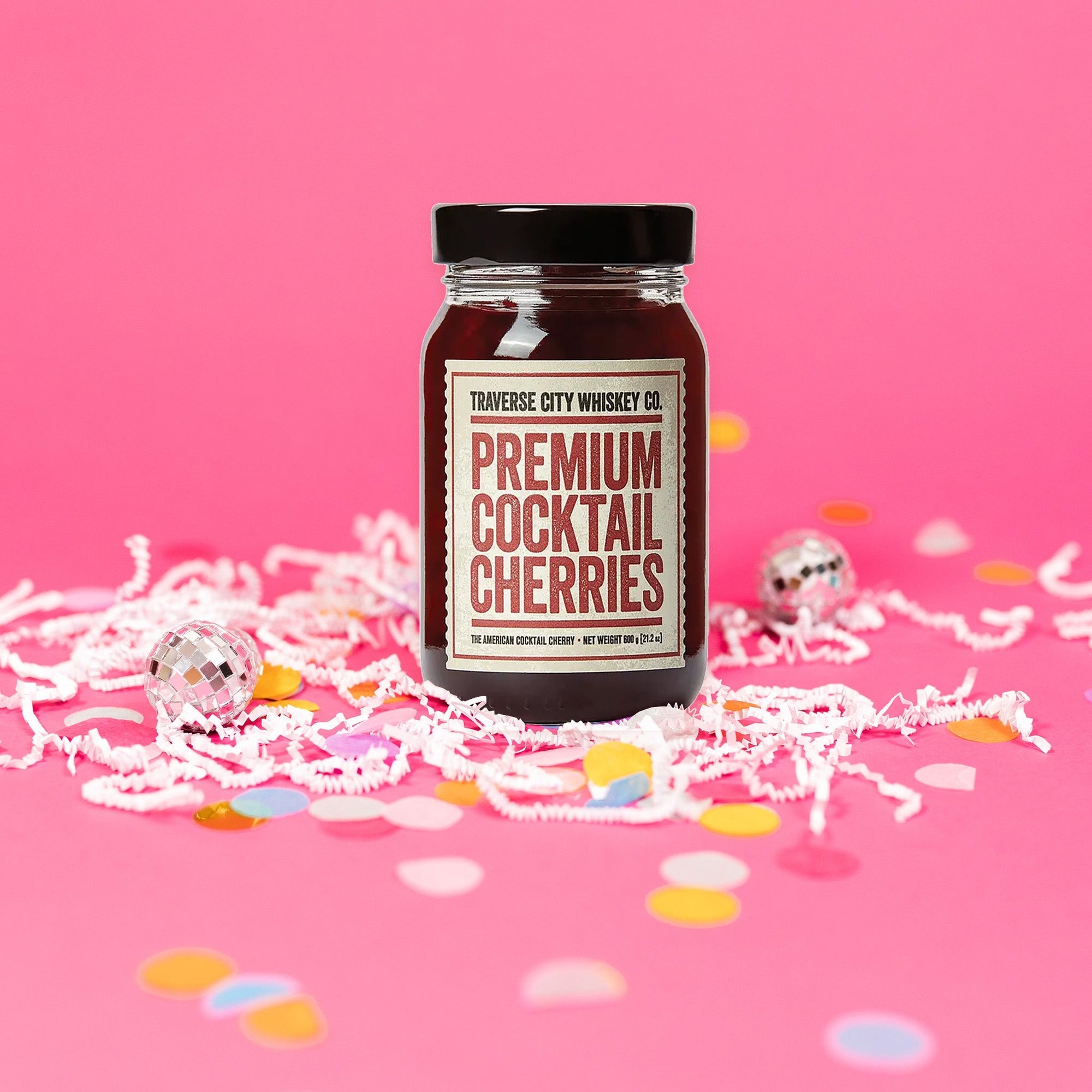 On a hot pink background sits a jar with white crinkle and big, colorful confetti. There are mini disco balls. The jar has a vintage creamy label that says "TRAVERSE CITY WHISKEY CO." in black, all caps block font. Under it is a red line and it says "PREMIUM COCKTAIL CHERRIES" in a red, all caps block font. There is another red line under that and then it says "THE AMERICAN COCKTAIL CHERRY • NET WEIGHT 600 g (21.2 oz)."
