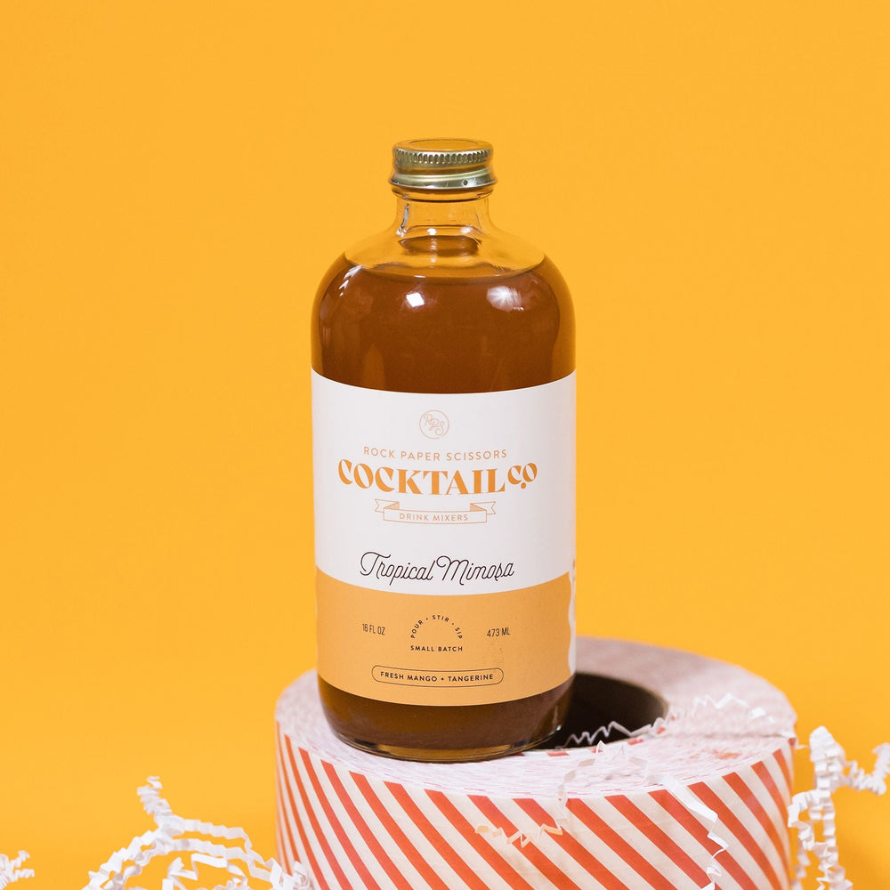 On a sunny mustard background sits a bottle and packing tape surrounded by white crinkle. The clear bottle is a Rock Paper Scissors Cocktail Co. Drink Mixer called "Tropical Mimosa". The flavors are fresh mango and tangerine. It has a white label with a mustard yellow stripe at the bottom. The bottle sits atop a red and white striped packing tape. 16 oz (473ml)