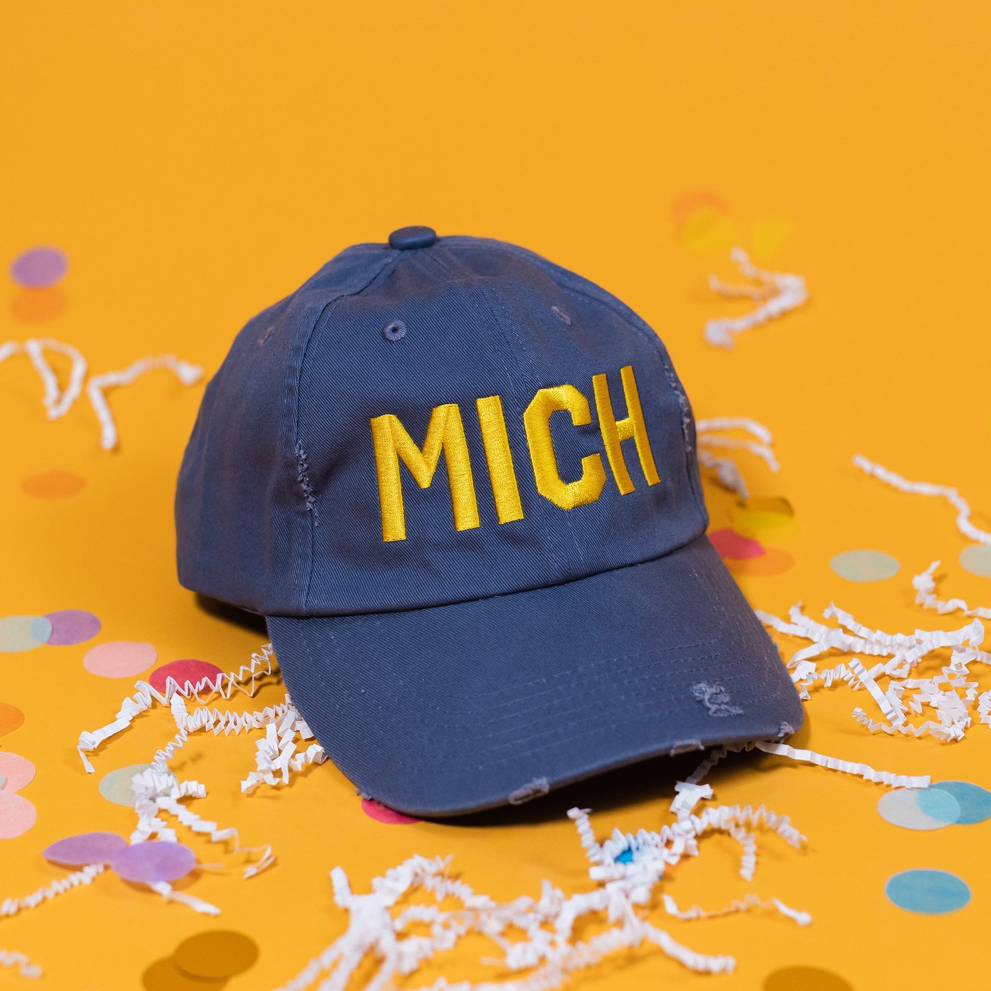 On a sunny mustard background sits a navy hat with white crinkle and big, colorful confetti scattered around. The hat is distressed with bold, yellow embroidered lettering that says "MICH."