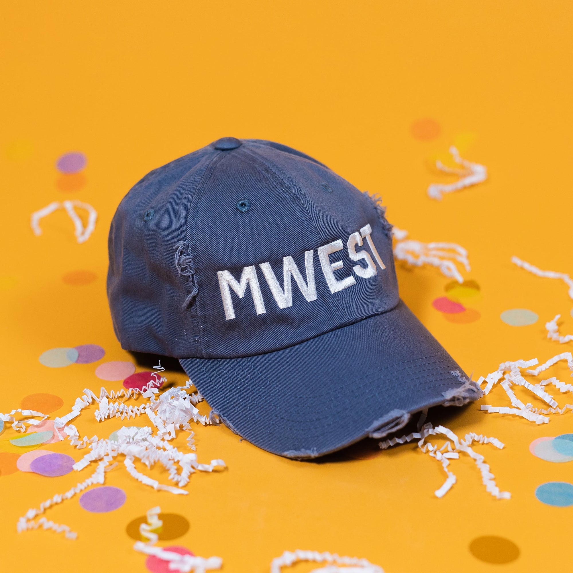 On a sunny mustard background sits a navy hat with white crinkle and big, colorful confetti scattered around. The hat is distressed with bold, white embroidered lettering that says "MWEST."