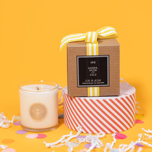 On a sunny mustard background is a candle with the back of a box and white crinkle with big, colorful confetti scattered around. The clear glass candle has a gold foil round label with an illustration of a bee on it. There is a kraft box with yellow and white striped ribbon tied at the top and on the back is a square black label that says "No. 8 Amber Musk and Sage, Made in the USA, 60+ Hour Burn."