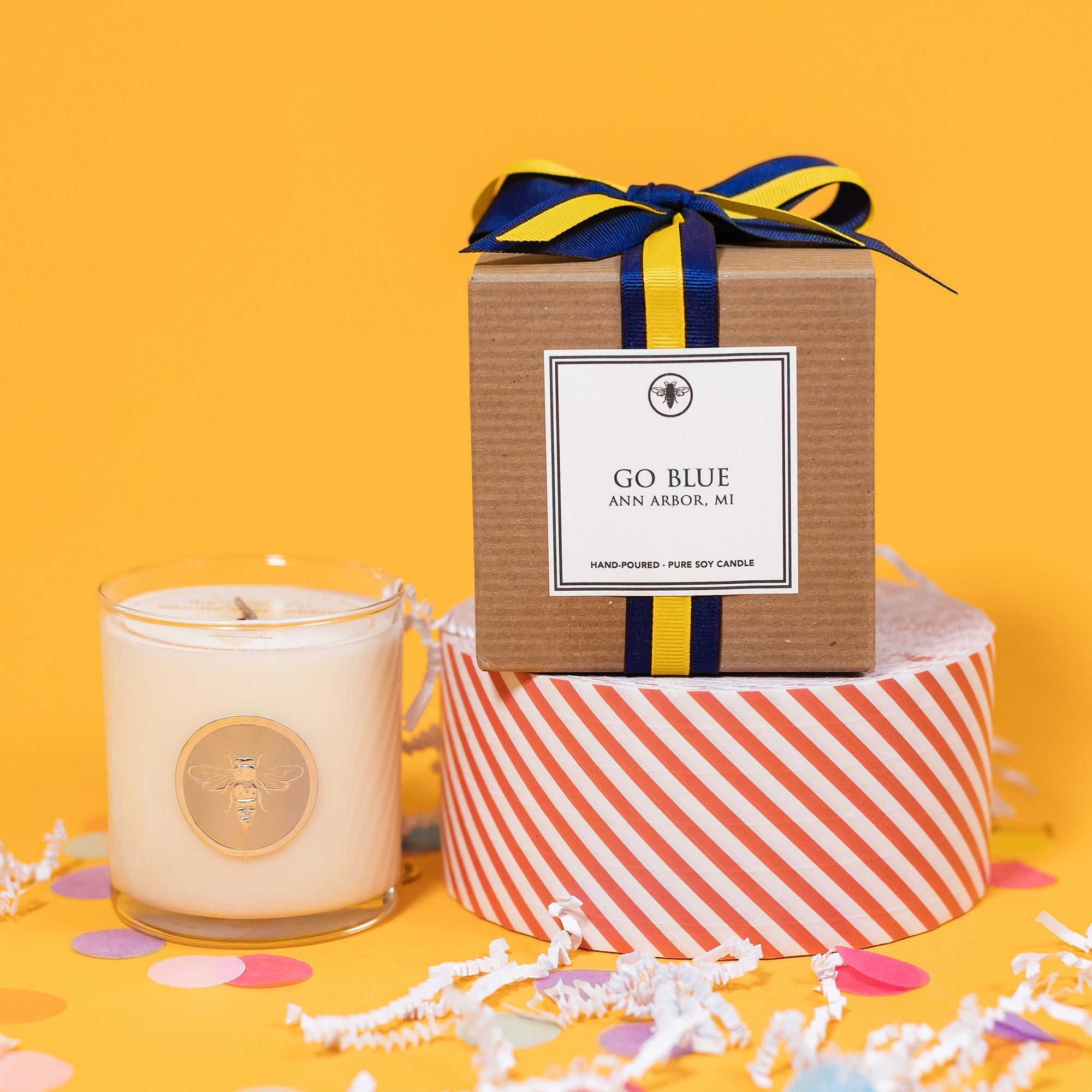 On a sunny mustard background is a candle with a box and white crinkle with big, colorful confetti scattered around. The clear glass candle has a gold foil round label with an illustration of a bee on it. There is a kraft box with yellow and navy striped ribbon tied at the top and on the front is a square white label that says "Go Blue Ann Arbor Mi" and it sits atop a red and white striped packing tape. The candle is a "Hand-poured, pure soy candle."