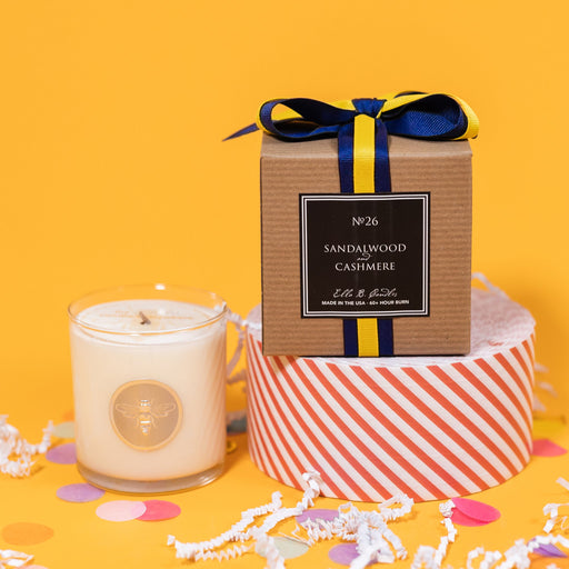 On a sunny mustard background is a candle with the back of a box and white crinkle with big, colorful confetti scattered around. The clear glass candle has a gold foil round label with an illustration of a bee on it. There is a kraft box with yellow and navy striped ribbon tied at the top and on the back is a square black label that says "No. 26 Sandalwood and Cashmere, Made in the USA, 60+ Hour Burn."