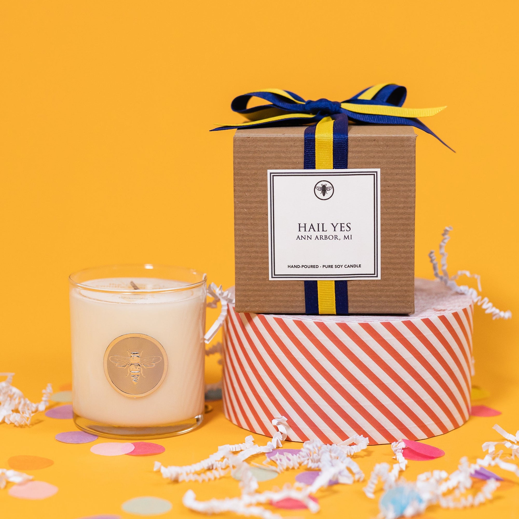 On a sunny mustard background is a candle with a box and white crinkle with big, colorful confetti scattered around. The clear glass candle has a gold foil round label with an illustration of a bee on it. There is a kraft box with yellow and navy striped ribbon tied at the top and on the front is a square white label that says "Hail Yes Ann Arbor Mi" and it sits atop a red and white striped packing tape. The candle is a "Hand-poured, pure soy candle."