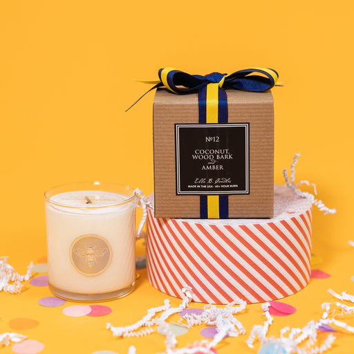 On a sunny mustard background is a candle with the back of a box and white crinkle with big, colorful confetti scattered around. The clear glass candle has a gold foil round label with an illustration of a bee on it. There is a kraft box with yellow and navy striped ribbon tied at the top and on the back is a square black label that says "No. 12 Coconut, Wood Bark and Amber, Made in the USA, 60+ Hour Burn."