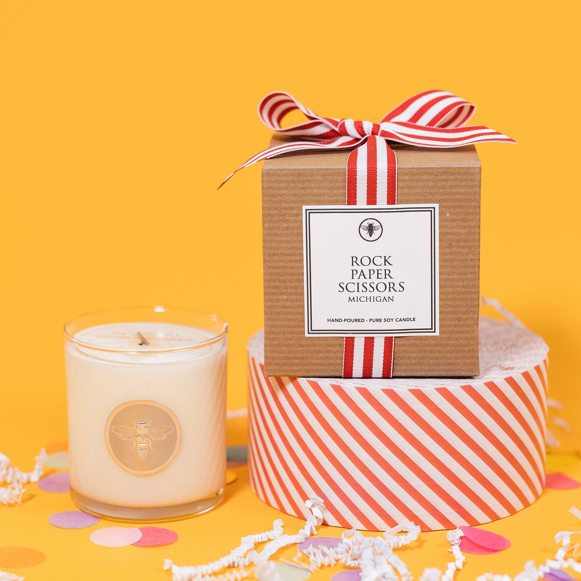 On a sunny mustard background is a candle with a box and white crinkle with big, colorful confetti scattered around. The clear glass candle has a gold foil round label with an illustration of a bee on it. There is a kraft box with red and white striped ribbon tied at the top and on the front is a square white label that says "Rock Paper Scissors Michigan" and it sits atop a red and white striped packing tape. The candle is a "Hand-poured, pure soy candle."