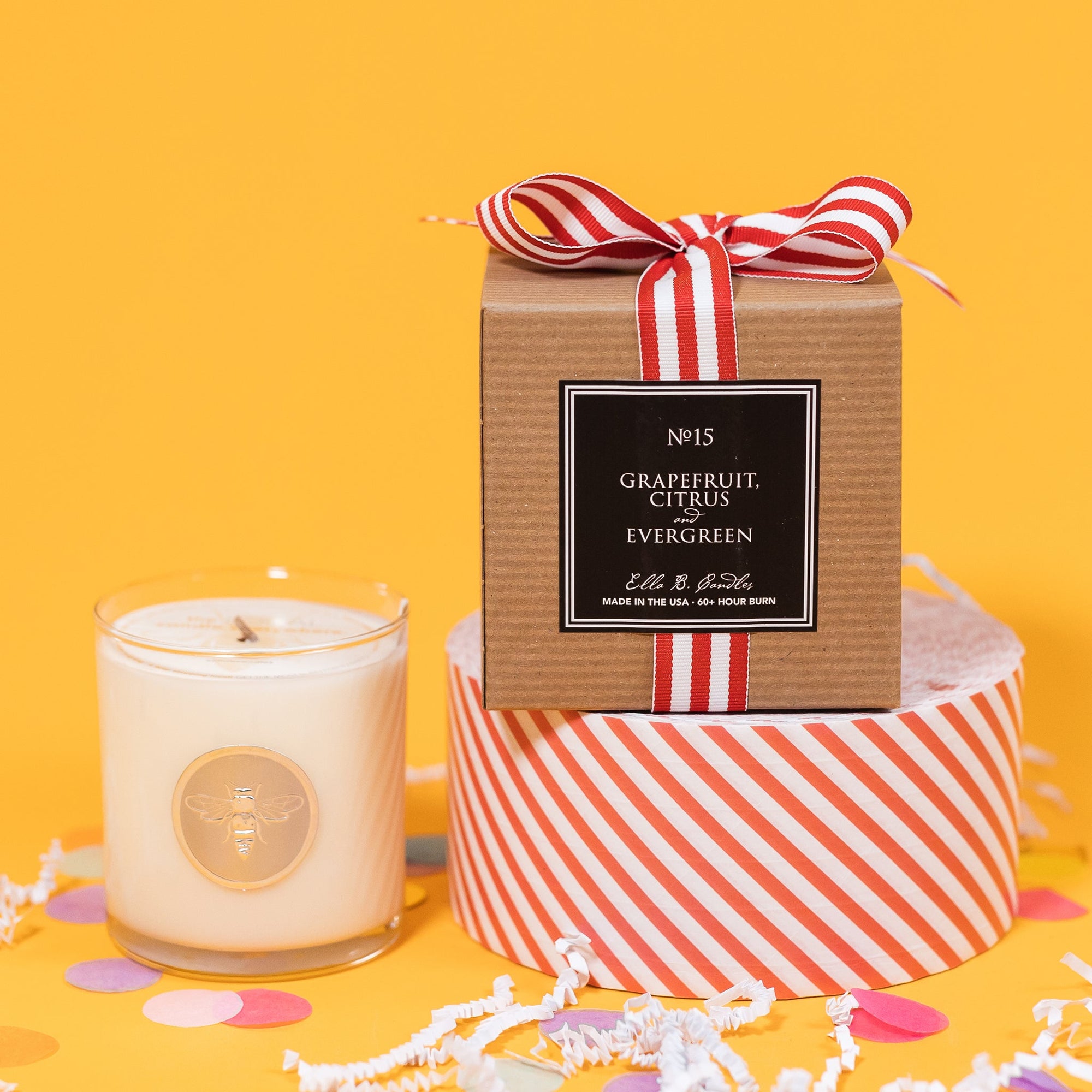 On a sunny mustard background is a candle with the back of a box and white crinkle with big, colorful confetti scattered around. The clear glass candle has a gold foil round label with an illustration of a bee on it. There is a kraft box with red and white striped ribbon tied at the top and on the back is a square black label that says "No. 15 Grapefruit, Citrus and Evergreen, Made in the USA, 60+ Hour Burn."