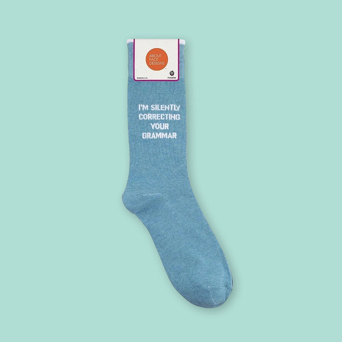 On a mint green background sits a sock. It is a light denim blue with white text on it. It says "I'M SILENTLY CORRECTING YOUR GRAMMAR" in all caps, block font.