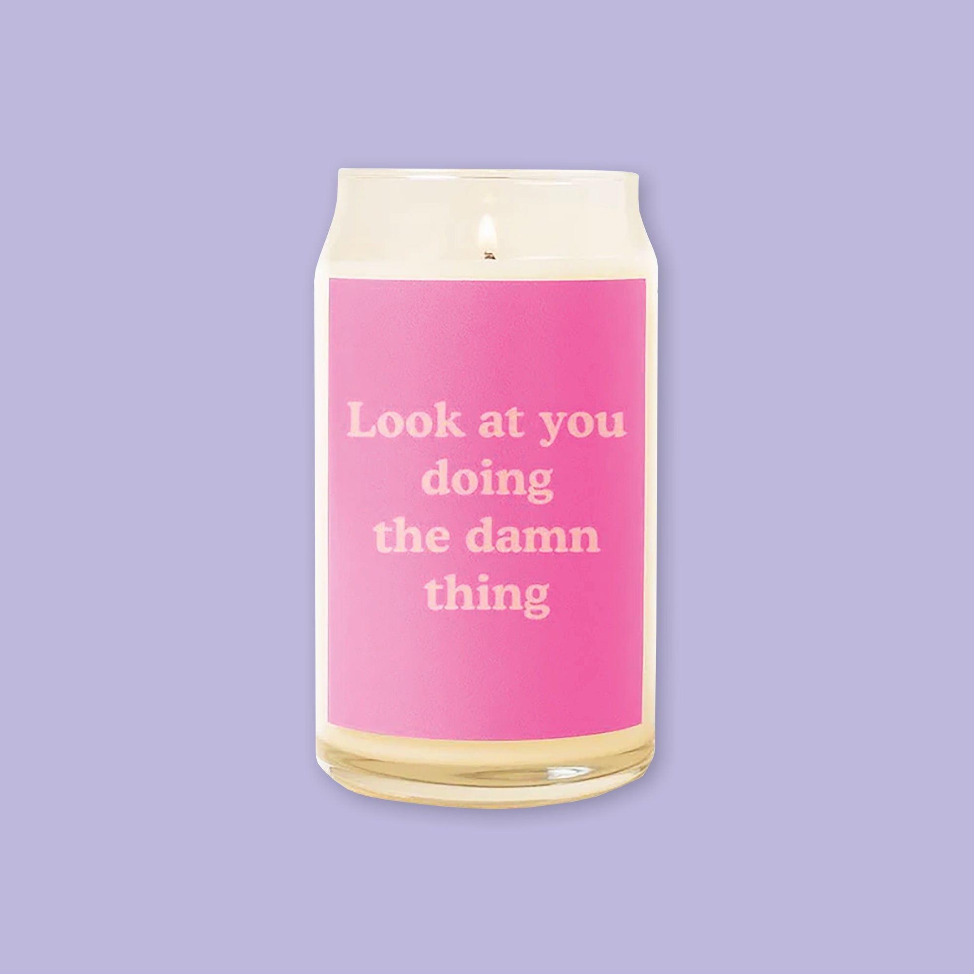 On a lavender background sits a candle. This beer can inspired glass candle is lit and has a hot pink label on the front. It says "Look at your doing the damn thing" in a coral, thick serif font.