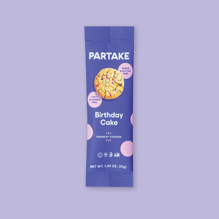 On a lavender background sits a package. This purple package has a picture of a cookie with colorful sprinkles on it and there are pink dots around it. It says "PARTAKE Birthday Cake" in white, block font. It also says "CRUNCHY COOKIES" in white, all caps block font. NET WT. 1.09 OZ. (31g)