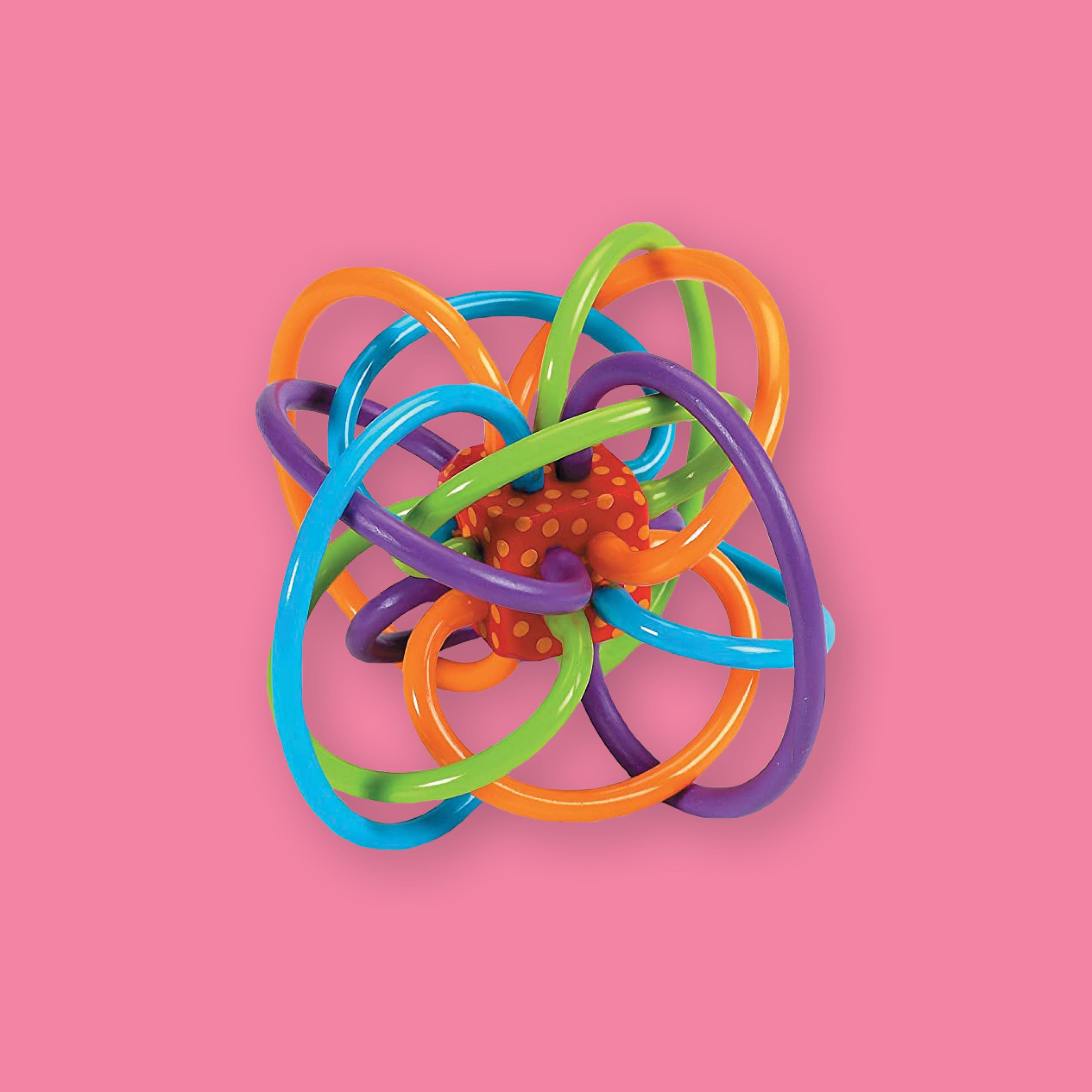 On a bubblegum pink background sits a teether rattle. This soothing Winkel teether rattle has colorful loops made from soft, pliable plastic. The colors are orange, bright blue, purple, and lime green. It has a red box in the center with orange dots covering it.