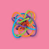 On a bubblegum pink background sits a teether rattle. This soothing Winkel teether rattle has colorful loops made from soft, pliable plastic. The colors are orange, bright blue, purple, and lime green. It has a red box in the center with orange dots covering it.