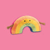 On a bubblegum pink background sits a rainbow squishy. This Amuseable Rainbow squishy has pastel stripes in peach, yellow and turquoise, cordy arms and a goofy smile.