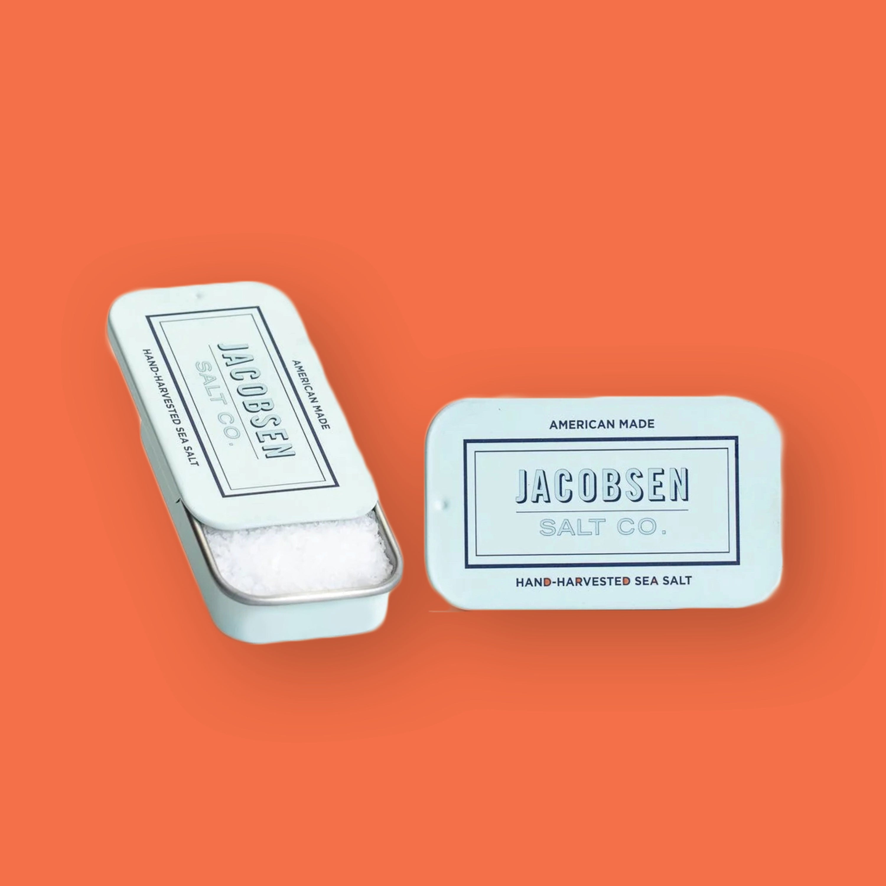 On an orangey-red background sits two rectangle tin cans. One tin can is open and shows sea salt sitting in it. The other shows the top of the tin can. Both are in a light seafoam blue color with navy writing and a rectangle. It says "AMERICAN MADE" in navy, all caps block font. It says "JACOBSEN SALT CO" in the rectangle, in navy outline, all caps block font. At the bottom it says "HAND-HARVESTED SEA SALT" in navy, all caps block font.