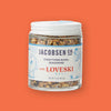 On an orangey-red background sits a jar. The clear jar has a white lid and label and shows various seasonings inside. It says "JACOBSEN CO. EVERYTHING BAGEL SEASONING" in navy, all caps block font. It says "with LOVESKI" in red font and "DELI" in a turquoise, all caps block font. There are illustrations in turquoise of a bagel with arms and a woman holding a bagel. New wt 2.2 oz (62.3g)
