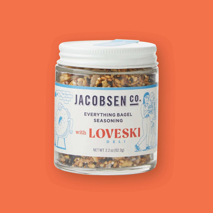 On an orangey-red background sits a jar. The clear jar has a white lid and label and shows various seasonings inside. It says "JACOBSEN CO. EVERYTHING BAGEL SEASONING" in navy, all caps block font. It says "with LOVESKI" in red font and "DELI" in a turquoise, all caps block font. There are illustrations in turquoise of a bagel with arms and a woman holding a bagel. New wt 2.2 oz (62.3g)