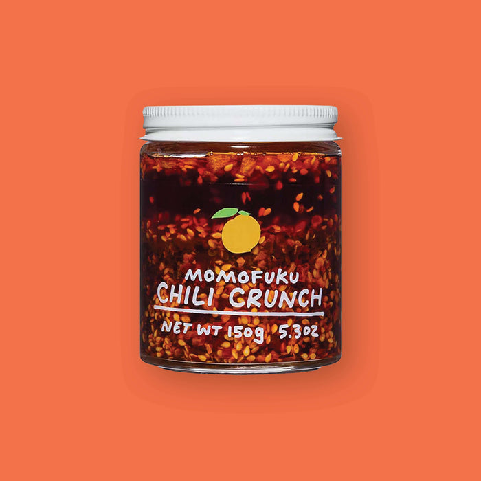On an orangey-red background sits a jar. This clear jar has a white lid and is filled with chili seeds and liquid. On the front is an illustration of a yellow lemon and it says in white "MOMOFUKU CHILI CRUNCH" underlined in handwritten lettering. Net wt 150g 5.3oz