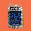 On an orangey-red background sits a package. This clear package has carmel popcorn in it with a black label. It says "SOUTH BEND CHOCOLATE COMPANY" in white, all caps block font and "SEA SALT CRUNCH" in a blue, all caps block font. 8 ounces | 227 grams