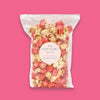On a hot pink background sits a package. This clear package is filled with pink and natural popcorn. There is a round, light pink label on the front that says "THE POPCORN SHOP" in all caps, font. It is in colors of purple, turquoise, lavender, light blue, hot pink, and mustard yellow. It says "GOURMET POPCORN" in purple, all caps font. There are two turquoise lines and in between that it says "STRAWBERRY SHORTCAKE" in purple, all caps font.
