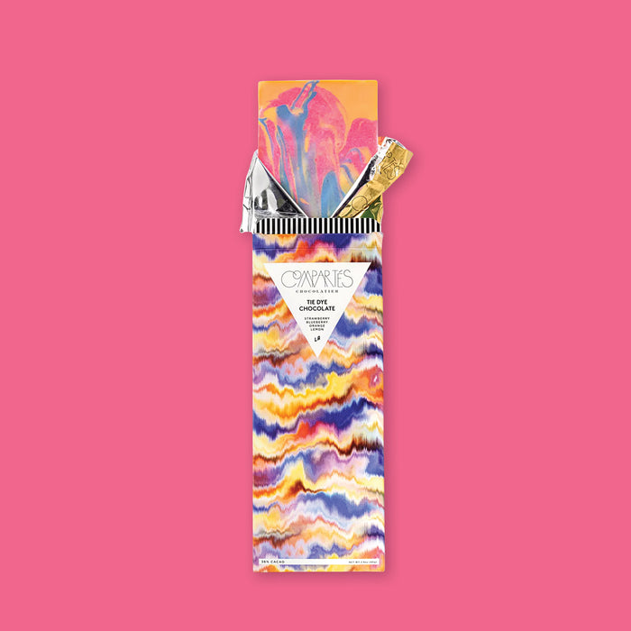 On a hot pink background sits an opened package. This colorful, tie dye package has a tie dye chocolate bar sticking out of the top with gold and silver foil that is opened. The colors of the tie dye are yellow mustard, hot pink, and purple. On the front of the package is a white triangle and it says "COMPARTES CHOCOLATIER," "TIE DYE CHOCOLATE," "STRAWBERRY BLUEBERRY ORANGE LEMON," "LA" all in black font. 36% CACAO NET WT 3OZ (85G)