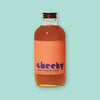 On a mint background sits a bottle. This clear bottle has a golden amber liquid in it and an orange label on the front. It says "cheeky" in a purple, lower case block font. It also says "HONEY GINGER SYRUP" in purple, all caps block font. Shake well • Shelf-Stable • 4 Fl Oz