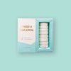 On a mint background sits two boxes. This picture is a close-up of a white and pool blue package that says "I NEED A VACATION" in gold foil, all caps block lettering. Under it says "set of 8" and " shower steamers" in grey, lowercase serif font. At the bottom it says "coconut lime" in gold foil, lower case serif font. To the right of it is a pool blue box with white shower steamers in it.