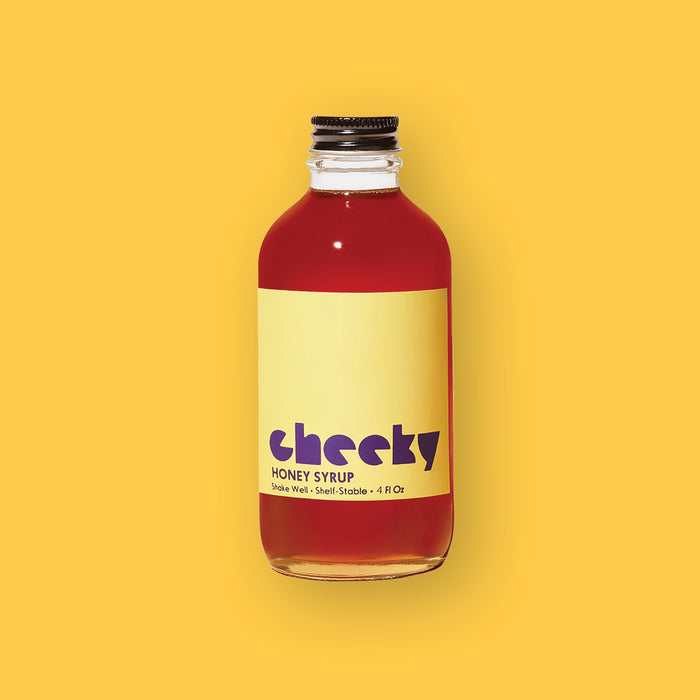 On a sunny mustard background sits a bottle. This clear bottle has a golden amber liquid in it and a yellow label on the front. It says "cheeky" in a purple, lower case block font. It also says "HONEY SYRUP" in purple, all caps block font. Shake well • Shelf-Stable • 4 Fl Oz