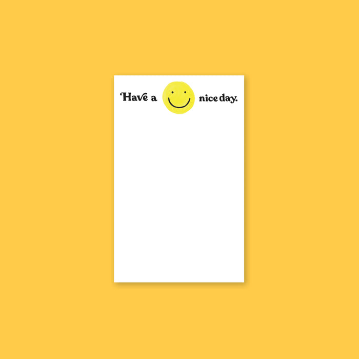 On a sunny mustard background sits a white notepad with a handdrawn yellow, smily face. It says "Have a nice day." in black, handwritten lettering.