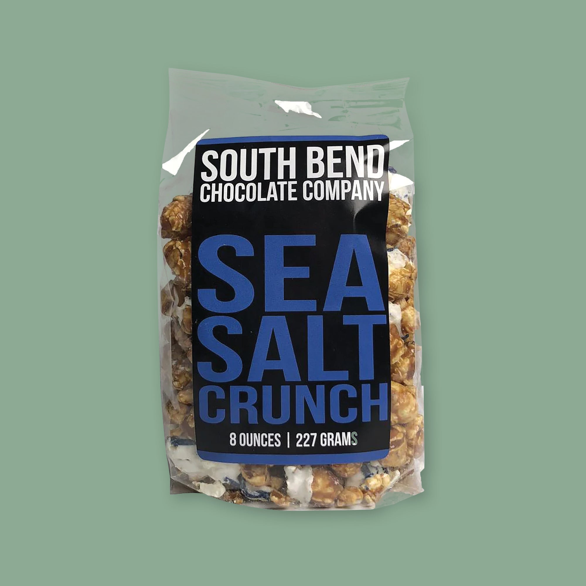 On a moss green background sits a package. This clear package is full of caramel popcorn. It has a black label on the front and it says "SOUTH BEND CHOCOLATE COMPANY" in white, all caps block font. It also says in a bright blue "SEA SALT CRUNCH" in all caps, block font. 8 OUNCES | 227 GRAMS