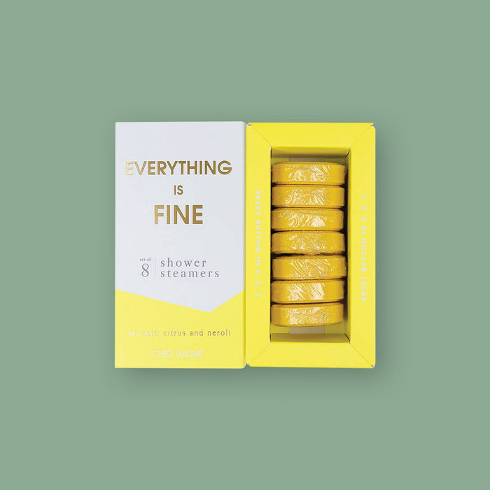 On a moss green background sits two boxes. This picture is a close-up of a white and yellow package that says "EVERYTHING IS FINE" in gold foil, all caps block lettering. Under it says "set of 8" and " shower steamers" in grey, lowercase serif font. At the bottom it says "sea salt citrus and neroli" in gold foil, lower case serif font. To the right of it is a yellow box with yellow shower steamers in it.