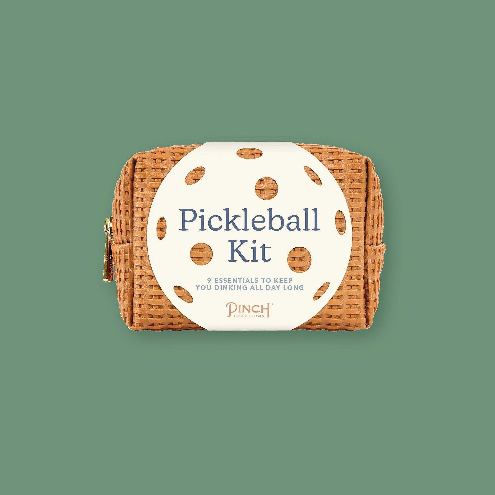 On a moss green background sits a kit. This light brown woven kit has a cream, round label with holes on it that looks like a ball. It says "Pickleball Kit" in a blue, serif font. Under that it says "9 ESSENTIALS TO KEEP YOU DINKING ALL DAY LONG" in light blue, all caps block font. It also says "PINCH PROVISIONS" in light brown, all caps font.