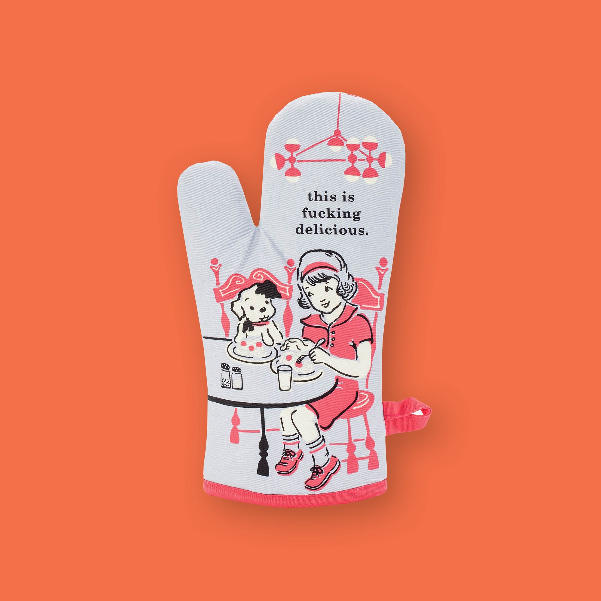 On an orangey-red background sits an oven mitt. This oven mitt has a vintage style illustration of a young girl sitting at a table eating spaghetti wiht her black and white dog. The colors are in light red, black, and white. It says "this is fucking delicious." in black, serif font.