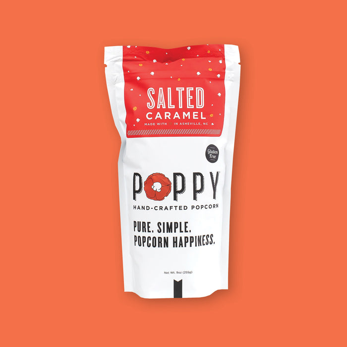 On an orangey-red background sits a white package. This package has a red label at the top with illustrations of white popcorn and orange carmel. It says "SALTED CARAMEL" in white, all caps font. It also says "MADE WITH (A RED HEART) IN ASHEVILLE, NC" in white, all caps font. It is gluten free. It says "POPPY HAND-CRAFTED POPCORN" and "PURE. SIMPLE. POPCORN HAPPINESS." in black, all caps font. New Wt. 9oz (255g)