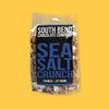 On a sunny mustard background sits a package. This clear package is full of caramel popcorn. It has a black label on the front and it says "SOUTH BEND CHOCOLATE COMPANY" in white, all caps block font. It also says in a bright blue "SEA SALT CRUNCH" in all caps, block font. 8 OUNCES | 227 GRAMS