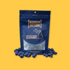 On a sunny mustard background sits a navy package that is see-through at the bottom. It is filled with chocolate covered blueberries. It says "MICHIGAN AWESOME," and "CHOCOLATE COVERED BLUEBERRIES" in goldish-yellow, all caps block font. It has an outlined illustration of the state of Michigan-UP.There are chocolate covered blueberries sitting around the bag. NET WT. 8 OZ. (227G)