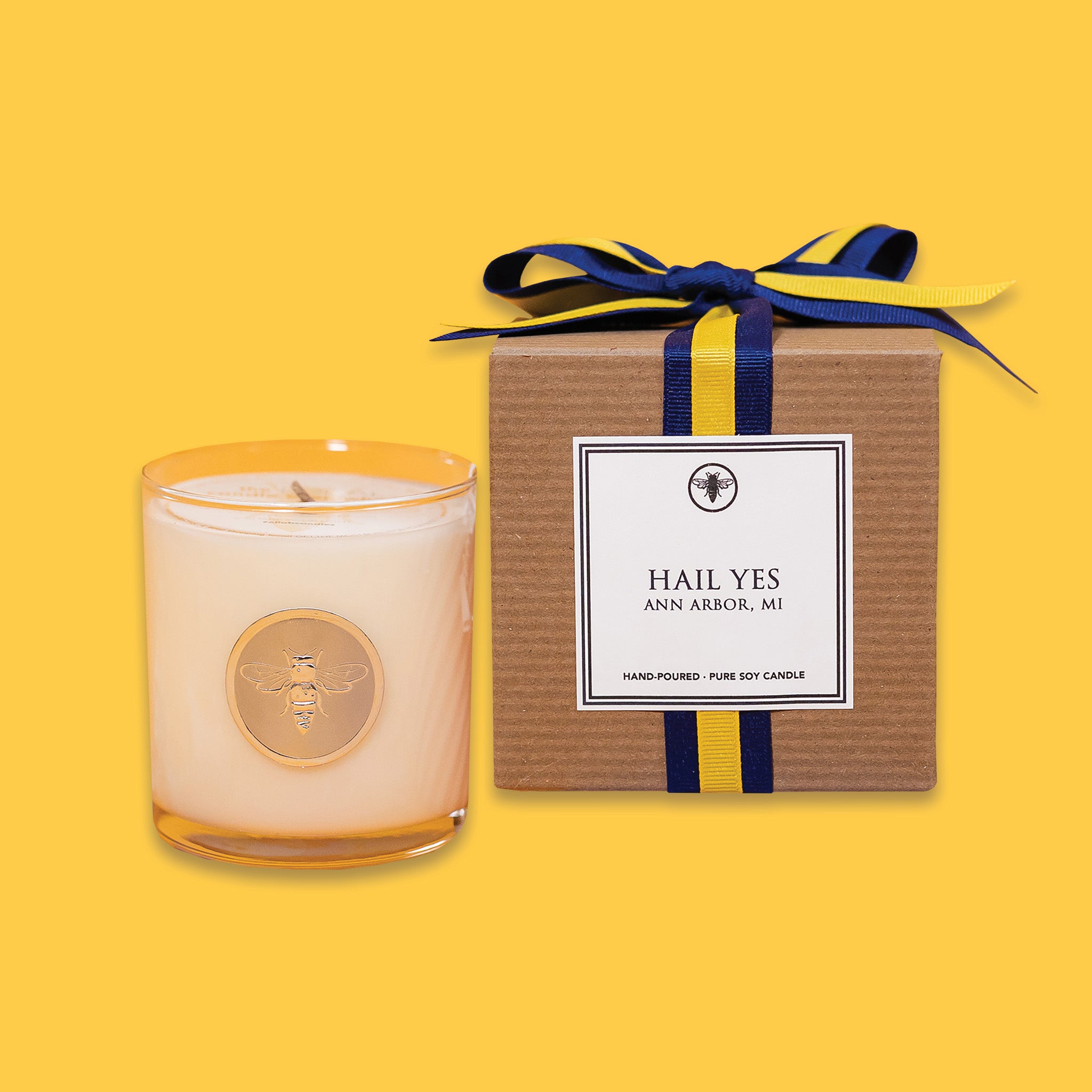 On a sunny mustard background is a candle with a box. The clear glass candle has a gold foil round label with an illustration of a bee on it. There is a kraft box with yellow and navy striped ribbon tied at the top and on the front is a square white label that says "HAIL YES ANN ARBOR, MI" in black, all caps serif font. The candle is a "Hand-poured, pure soy candle."