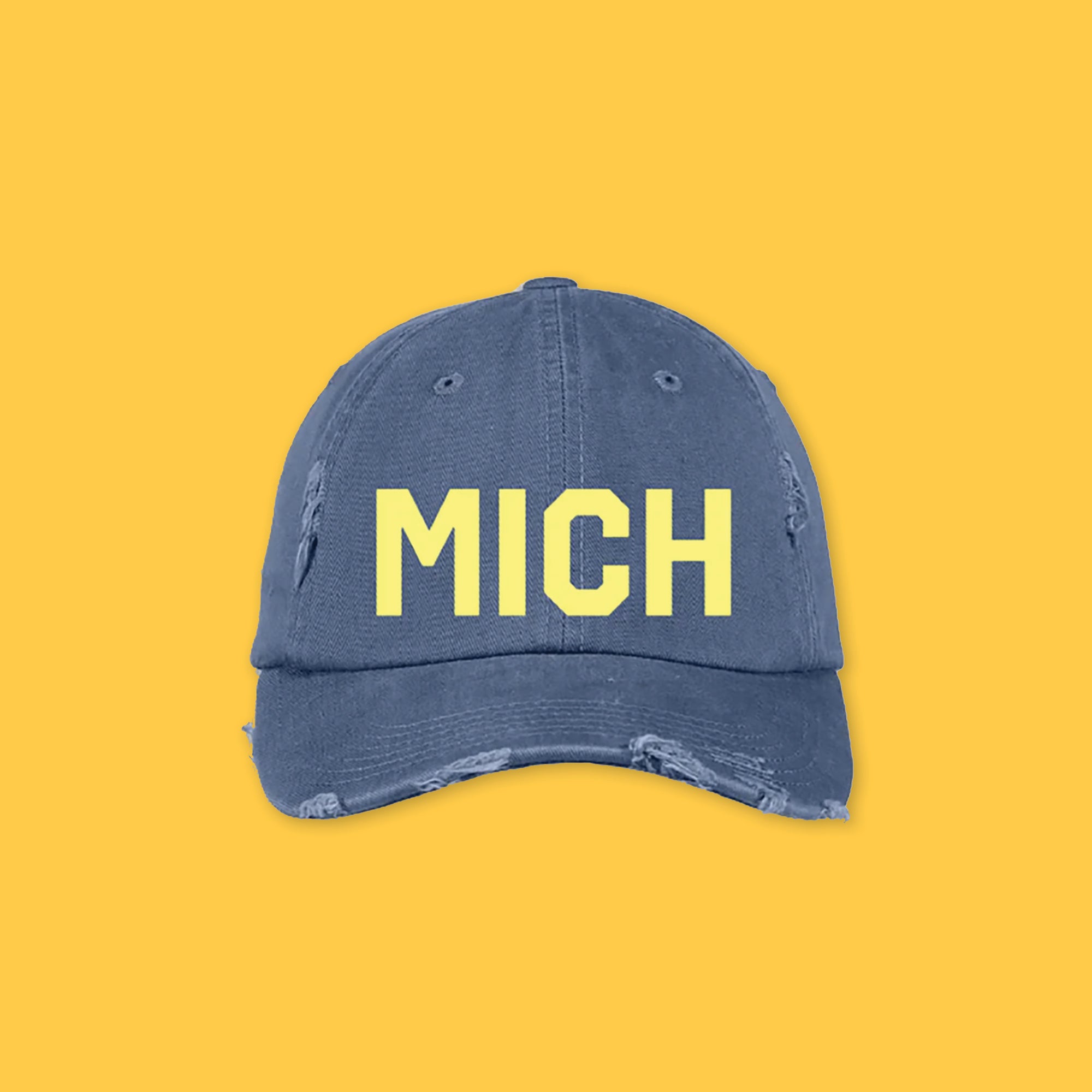 On a sunny mustard background sits a navy hat. The hat is distressed with bold, yellow embroidered lettering that says "MICH."
