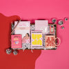 On a bright pink and red background, an assortment of Lizzo items are arranged among white packing crinkle and  mini disco balls. The gifts include a rose gold retractable gel pen, a notepad that says "It's About Damn Time" with an illustration of Lizzo, a bright pink book of morning affirmations, an Up and Up Salted Caramel Milk Chocolate bar, a mug that says "Be the Girl who Decided to Effing Go For It," gum that says, "Bitch I am the Secret Ingredient" and a set of Patchology 'Happy Place' eye masks.