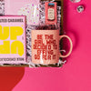 On a bright pink and red background, an assortment of Lizzo items are arranged among white packing crinkle and  mini disco balls. The photo is a close-up of an Up and Up Salted Caramel Milk Chocolate bar, a mug that says "Be the Girl who Decided to Effing Go For It," gum that says, "Bitch I am the Secret Ingredient" and a set of Patchology 'Happy Place' eye masks.