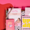 On a bright pink and red background, an assortment of Lizzo items are arranged among white packing crinkle and  mini disco balls. The photo is a close-up of a rose gold retractable gel pen, a notepad that says "It's About Damn Time" in pink with an illustration of Lizzo, a bright pink book of morning affirmations, an Up and Up Salted Caramel Milk Chocolate bar, and a set of Patchology 'Happy Place' eye masks.
