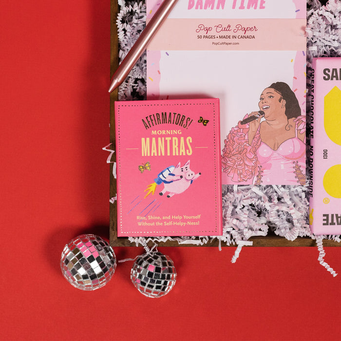 On a red background, an assortment of Lizzo items are arranged among white packing crinkle and  mini disco balls. The products in the photo is a close up of a bright pink book of morning affirmations, a rose gold retractable gel pen and a notepad that says "It's About Damn Time" in pink with an illustration of Lizzo.