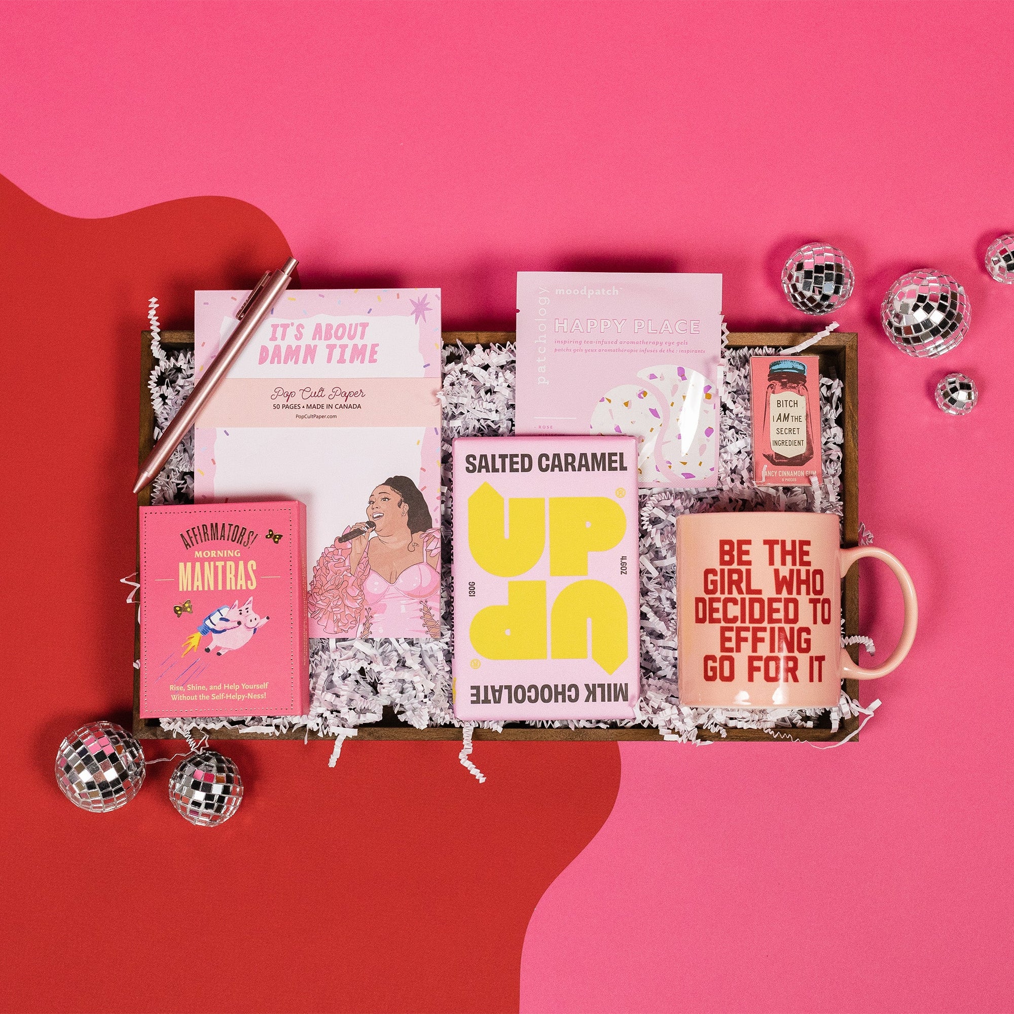 On a bright pink and red background, an assortment of Lizzo items are arranged among white packing crinkle and  mini disco balls. The gifts include a rose gold retractable gel pen, a notepad that says "It's About Damn Time" with an illustration of Lizzo, a bright pink book of morning affirmations, an Up and Up Salted Caramel Milk Chocolate bar, a mug that says "Be the Girl who Decided to Effing Go For It," gum that says, "Bitch I am the Secret Ingredient" and a set of Patchology 'Happy Place' eye masks.