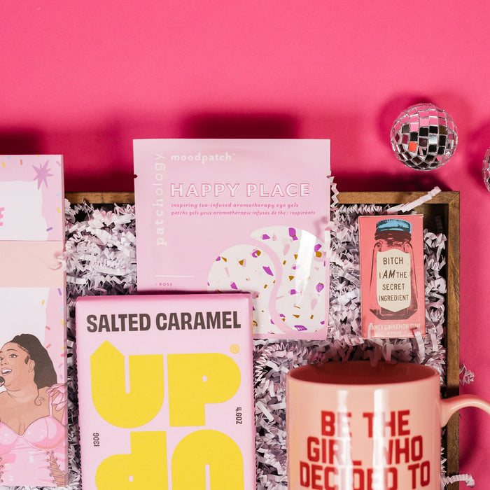 On a bright pink background, an assortment of Lizzo items are arranged among white packing crinkle and  mini disco balls. The photo is a close-up of a mug that says "Be the Girl who Decided to Effing Go For It" in red collegiate font, an Up and Up Salted Caramel Milk Chocolate bar, gum that says "Bitch I am the Secret Ingredient" and a set of Patchology 'Happy Place' eye masks.
