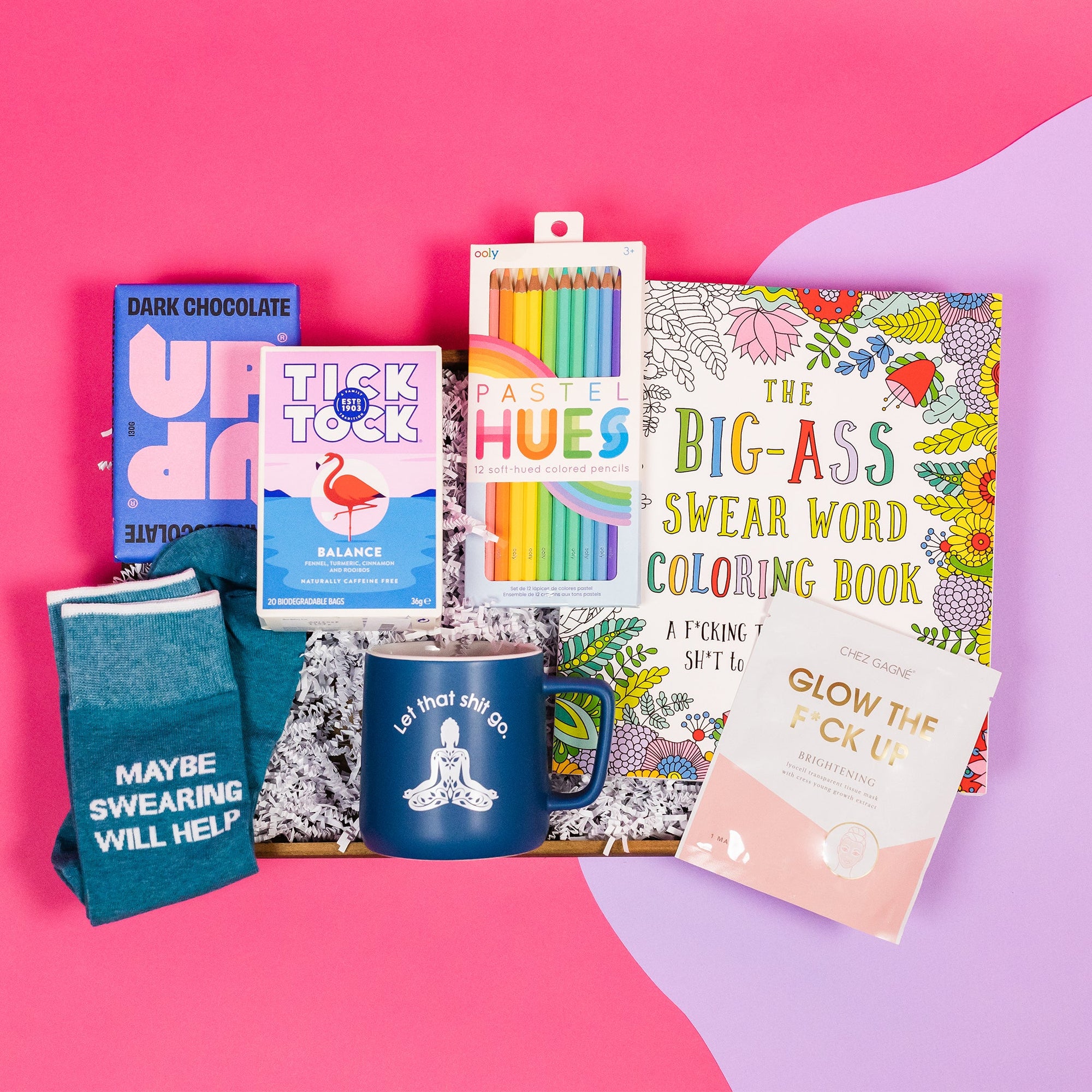 On a bright pink and purple background, a handful of optimistic, swear-themed gifts are arranged on a tray among white crinkle. The gifts includes a navy mug with a buddha sillhouette that says "Let that shit go," Tick Tock Tea 'Balance' blend, Up and Up Dark chocolate bar, a pair of blue novelty socks that says "MAYBE SWEARING WILL HELP" in white, a Chez Gagne 'Glow the Fuck Up' sheet mask, a 'Big-Ass Swear Word Coloring Book' with flowers and Ooly Pastel hues colored pencils.