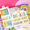 On a bright pink and purple background, a handful of optimistic, brightly colored swear-themed gifts are arranged on a tray among white crinkle. The photo is a close-up of the 'Big-Ass Swear Word Coloring Book' with flowers and Ooly Pastel hues colored pencils.