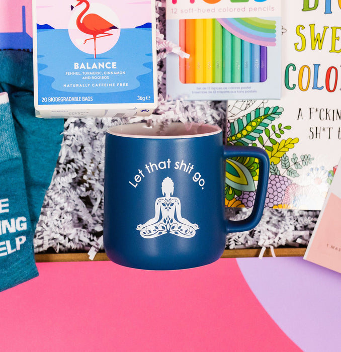 On a bright pink and purple background, a handful of optimistic, brightly colored swear-themed gifts are arranged on a tray among white crinkle. The photo is a closeup of the navy blue mug with white text and a buddha sillhouette that says "Let that shit go."