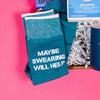 On a bright pink and purple background, a handful of optimistic, brightly colored swear-themed gifts are arranged on a tray among white crinkle. The photo is a close-up of the blue socks that say "MAYBE SWEARING WILL HELP" in white.