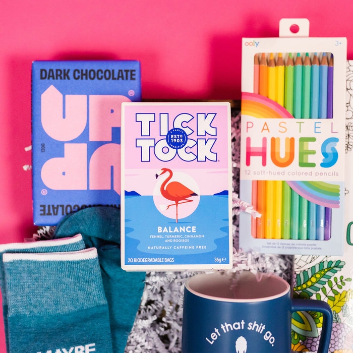 On a bright pink and purple background, a handful of optimistic, brightly colored swear-themed gifts are arranged on a tray among white crinkle. The photo is a close-up of the Tick Tock Tea Balance Blend, Up and UP Dark Chocolate bar and Ooly Pastel Hues colored pencils.