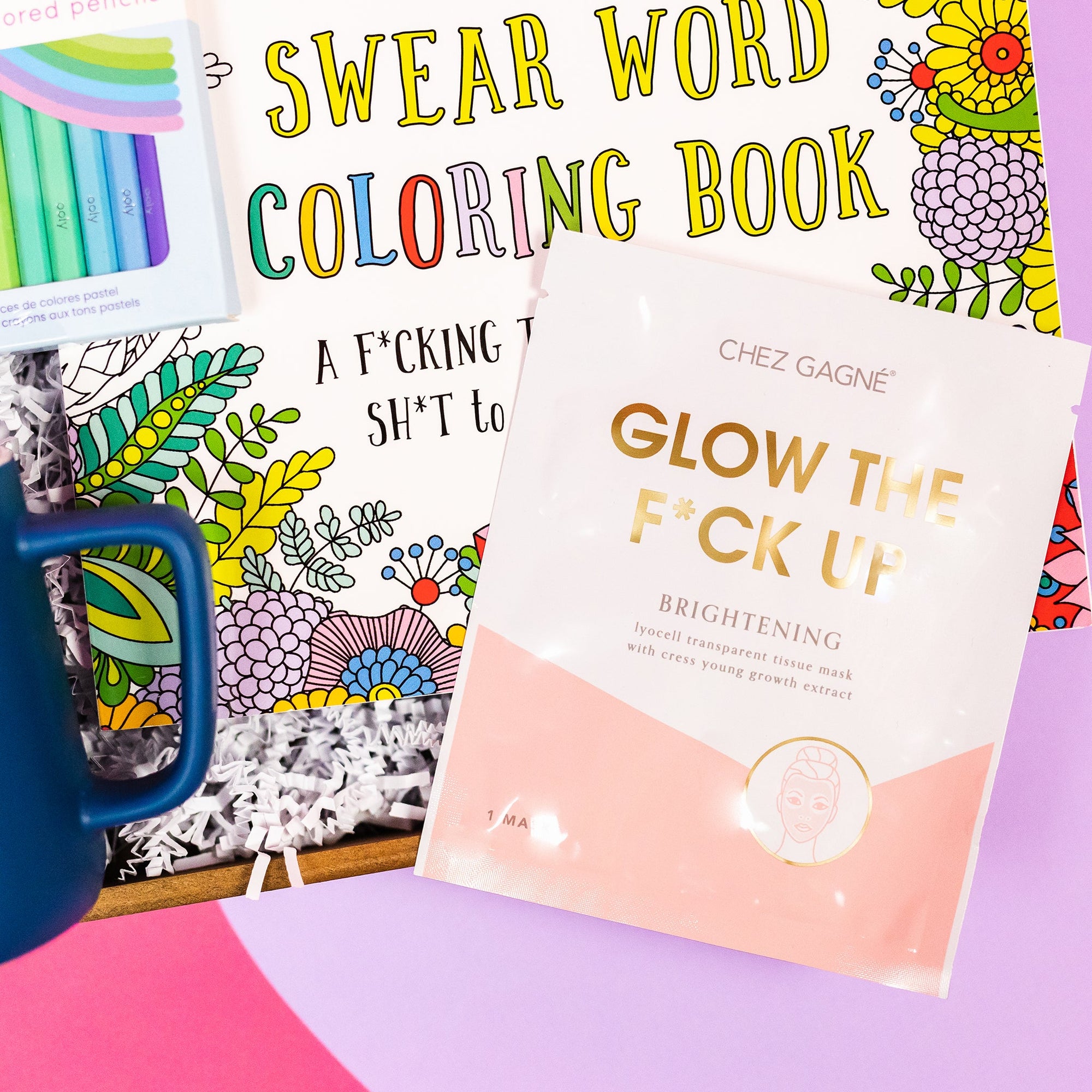 On a bright pink and purple background, a handful of optimistic, brightly colored swear-themed gifts are arranged on a tray among white crinkle. The photo is a close-up of the Chez Gagne Glow the Fuck Up Brightening face mask in a white and blush package with gold writing.