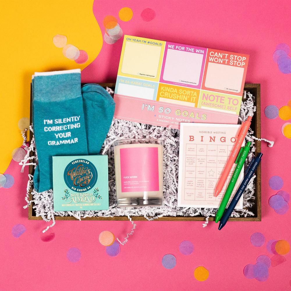 On a hot pink and yellow background, snarky, work-themed gifts are arranged in a tray among white paper crinkle with confetti. The gifts are a pack of "I'm So Goals" sticky notes, a pair of socks that say "I'M SILENTLY CORRECTING YOUR GRAMMAR," a candle that says "Fuck Work," Willie's Cacao Sea Kissed Almond Milk Chocolate, a Horrible Meeting Bingo pad, and a set of three colorful pens that say "Binged the Whole Season," "Hit Snooze at Least Twice," and "Ordered the Extra Large Coffee."